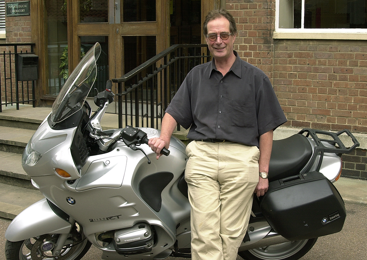 Dickinson is facing the camera leaning against a silver motorbike parked outside a brick building