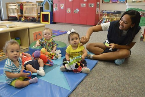 Three children are sitting on a blue and green mat; there are toys around them. A care giver is sitting next to the children and is trying to touch the cheek of one of the children.