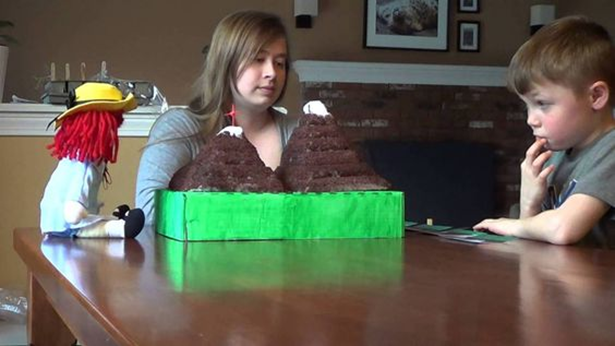 Woman watching a child who is sitting across from a child. In the center of the table is a model of two mountains.