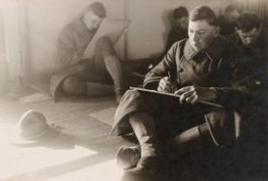 Group of people sitting on the floor indoors, each holding a drawing board and completing a test. A hat is placed on the floor to the left, and natural light enters from an unseen source.