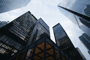 Low-angle view of several tall skyscrapers with modern glass and steel architecture, converging toward the sky. The lighting suggests early morning or late afternoon, with the sky visible between the buildings.