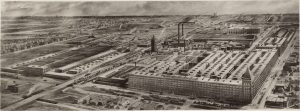 Aerial view of a large industrial complex featuring multiple buildings, smokestacks, and surrounding infrastructure. Roads, railways, and smaller buildings are visible nearby, with the landscape extending into the distance to include more structures and open land.