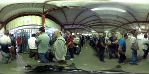 A large group of people standing in line inside a building with a corrugated metal roof. Metal barriers guide the orderly queue, suggesting a checkpoint or entrance area.
