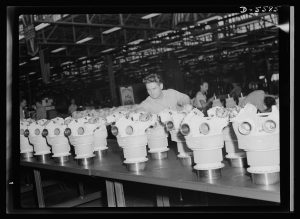 An industrial setting with multiple identical mechanical parts arranged in rows on a table. Several workers are in the background, and the environment appears to be a factory or assembly line. The mechanical parts have cylindrical bases with multiple circular openings on top. The image is black and white.