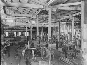 The interior of a large industrial workshop or factory with multiple workers engaged in various tasks. Machinery, tools, equipment, workbenches, and structural beams fill the space. The environment appears busy with several people focused on their work.