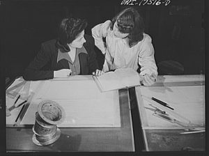 Two individuals working together at a desk. One is seated and reviewing a document, while the other stands and points to something on the paper. The desk contains drafting tools such as a ruler, pens, and a spool of wire or thread.