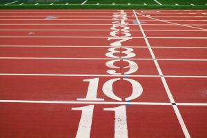 A section of a running track with lanes numbered from 1 to 11. The numbers are painted in white on the red surface of the track, and white lines separate each lane. The background includes a green field and additional track markings.