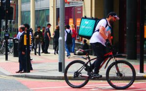Urban street scene with pedestrians walking near storefronts and a cyclist riding a bicycle. The cyclist wears a white t-shirt, black pants, and a black cap, and carries a large green 'Deliveroo' delivery bag. Several bicycles are parked at bike racks in the background.
