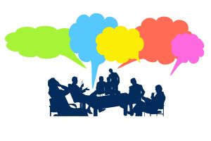 Silhouetted group of people seated around a table, actively engaged in discussion. Above them, colorful speech bubbles in green, blue, yellow, orange, and pink represent vibrant idea exchange and communication.