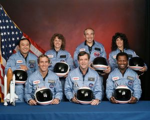Image of seven astronauts in blue NASA uniforms holding black helmets with white and red stripes. Four are standing in the back row, and three are seated in the front. An American flag is visible on the left side. The image is commonly associated with the Challenger disaster and used to highlight groupthink in organizational behavior.