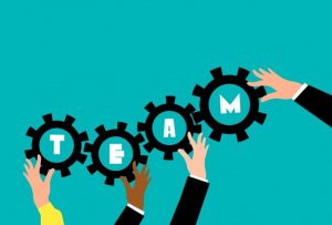 Four hands of varying skin tones and clothing styles hold black gears against a teal background. Each gear contains a white letter, collectively spelling the word TEAM, symbolizing diversity and collaboration.