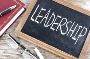 Small chalkboard with the word 'LEADERSHIP' written in white chalk, placed on a wooden surface alongside eyeglasses, an eraser, and a red notebook with a pen.