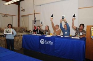 A panel of five judges seated behind a table with a "Conservation Through the Arts" banner, holding up scorecards—four showing the number 4 and one showing the number 5. To the left, a person stands holding up an artwork or photograph. The background features an American flag and decorative dried plants or reeds.