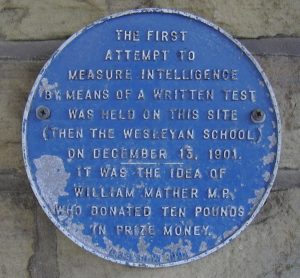 A blue circular plaque mounted on a wall with text commemorating the first attempt to measure intelligence by written test, held at the Wesleyan School on December 13, 1901, initiated by William Mather M.P., who donated ten pounds in prize money.