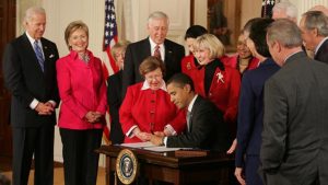 Group of people gathered around a man seated at a desk with the seal of the President of the United States, signing a document.