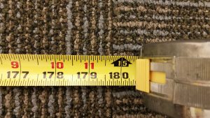 Close-up of a yellow measuring tape on a textured carpet, showing centimeter measurements from 177 to 180. Red numbers 9, 10, and 11 are marked above the corresponding centimeter marks, with an arrow pointing to the number 15 above 180.