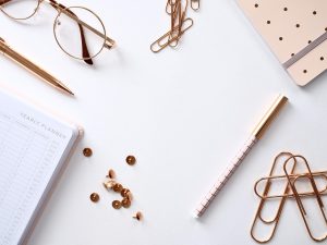 Flat lay of office supplies on a white surface, including round glasses, a gold pen, gold paper clips, a polka dot notebook with an elastic band, a yearly planner labeled "YEARLY PLANNER," scattered push pins, and a checkered pen.